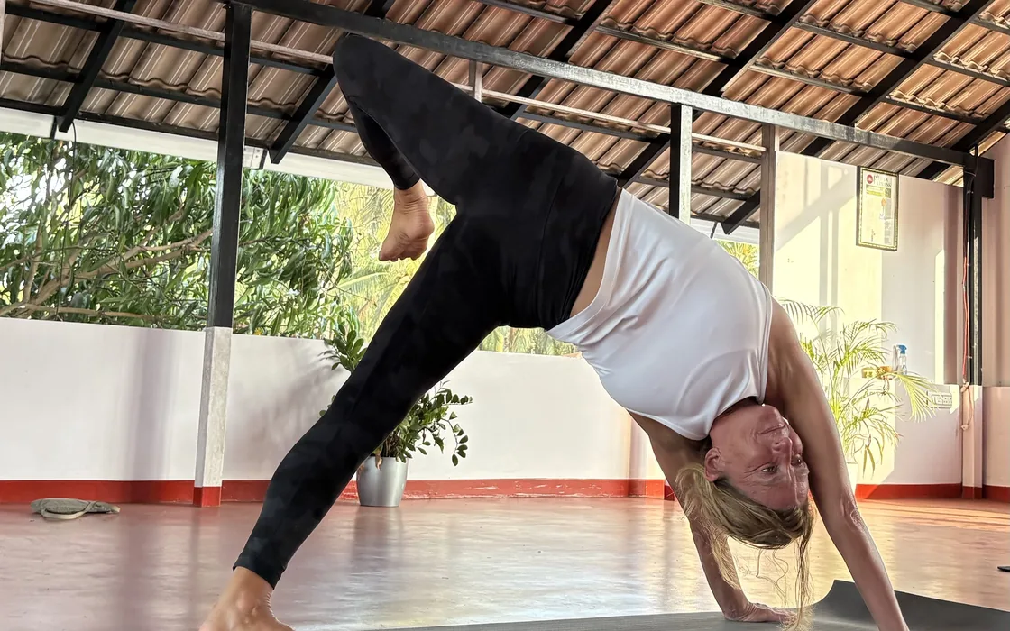 A student in a three-legged downward dog on the mat in the open-sided shala, one leg raised high.