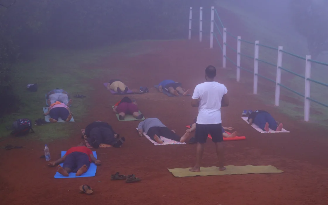 The retreat group in child's pose on the red earth of a misty hilltop at Matheran, Abhilash standing over them.