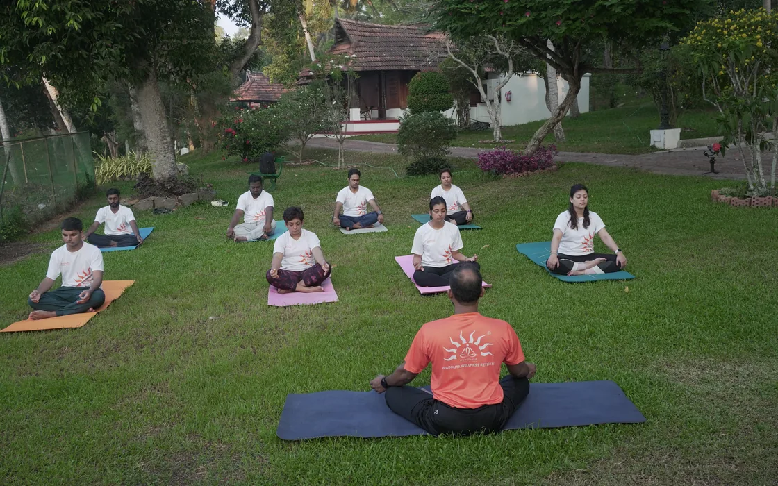 The retreat group seated in meditation on the lawn among the heritage cottages, Abhilash facing them.