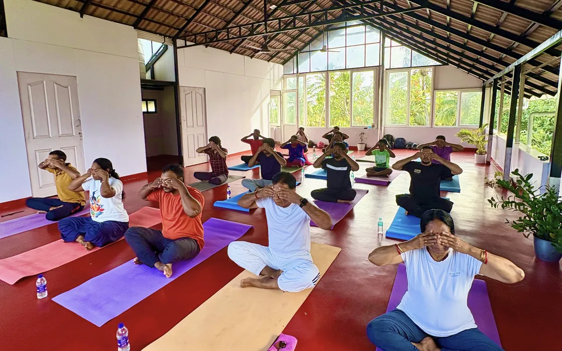 The International Day of Yoga class seated in the shala, hands over their faces in a breathing exercise.